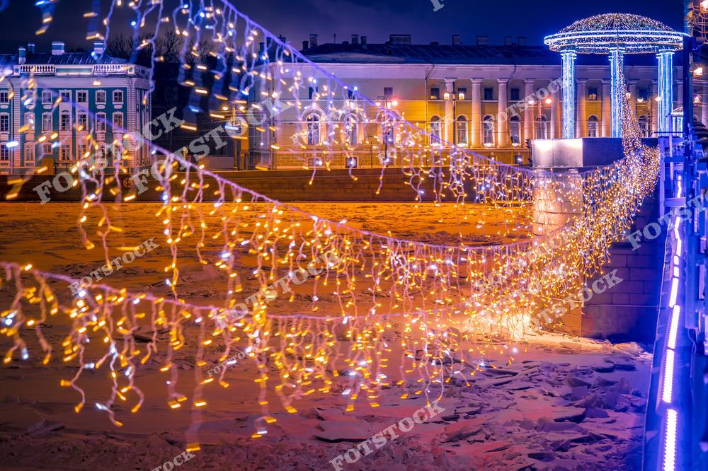 На переднем плане изображены новогодние гирлянды, украшающие мост, который соединяет берега замерзшей Невы. На заднем плане светящаяся ротонда и исторические здания Петербурга, подсвеченные ночной иллюминацией. Фотография передает атмосферу праздника и волшебства зимнего Санкт-Петербурга.
